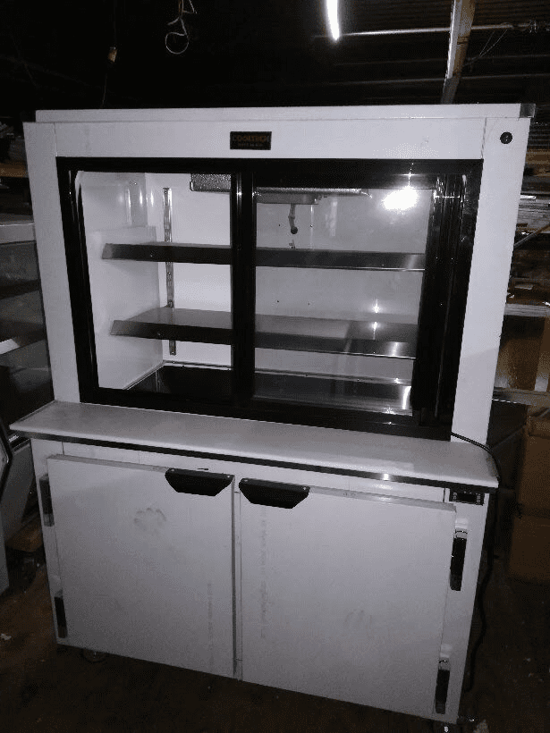 An empty Cooltech Pass-Through Display Pie Case Refrigerator 48” with sliding glass doors on top and closed cabinet doors below, stored in a dimly lit room.